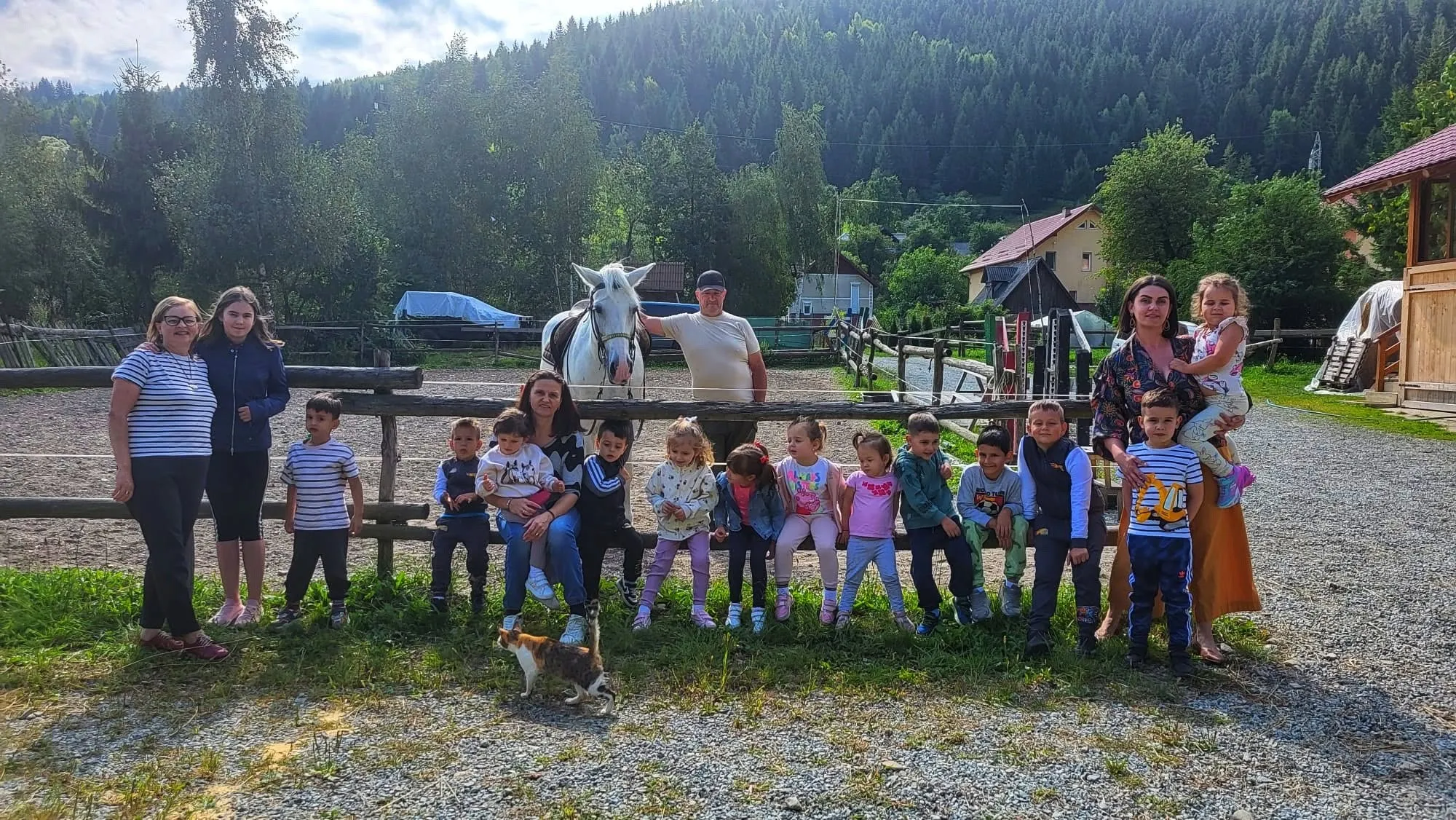 Un grup de copilași în vizită, alături de câțiva adulți, în fața padocului, cu un cal alb în spate
