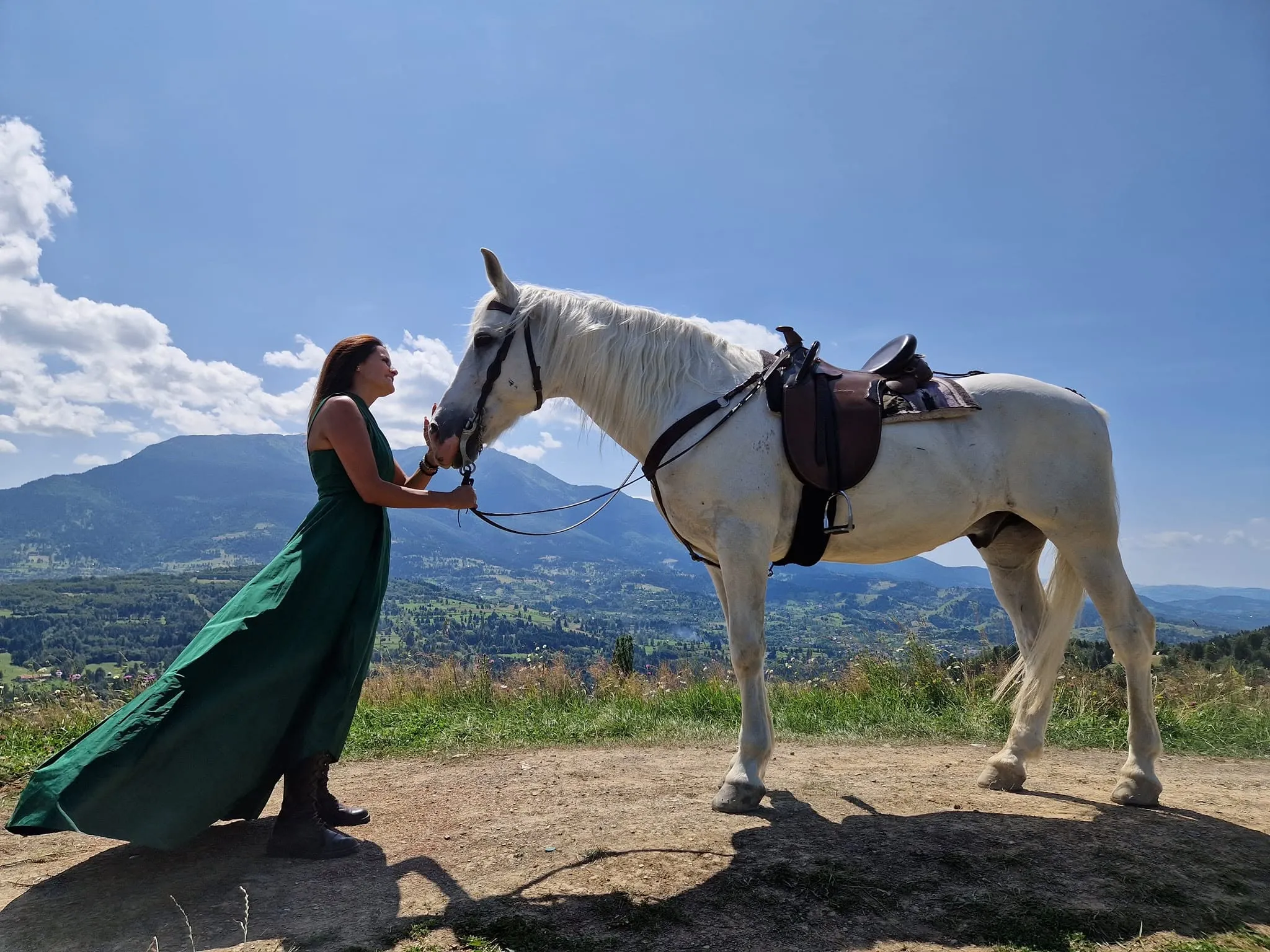 Mădălina, în rochie lungă verde, ține de căpăstru un cal alb înșeuat, pe un deal cu munți în fundal